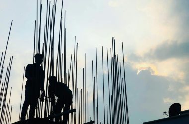 Silhouetted construction workers on site at dawn in Dhaka, Bangladesh, against a cloudy sky.